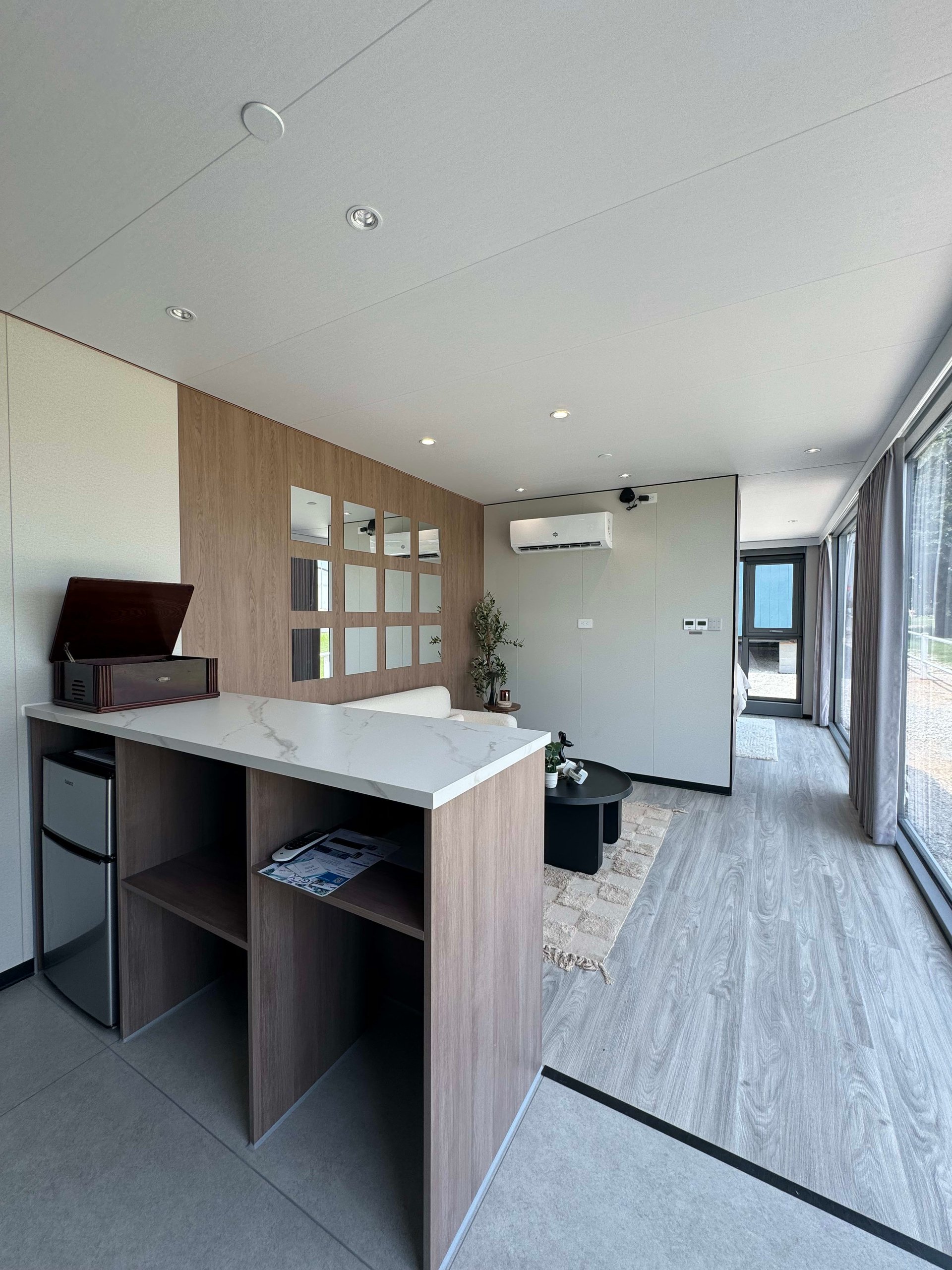Tiny home pod interior with a white counter, wood-paneled wall, compact refrigerator, and floor-to-ceiling windows.