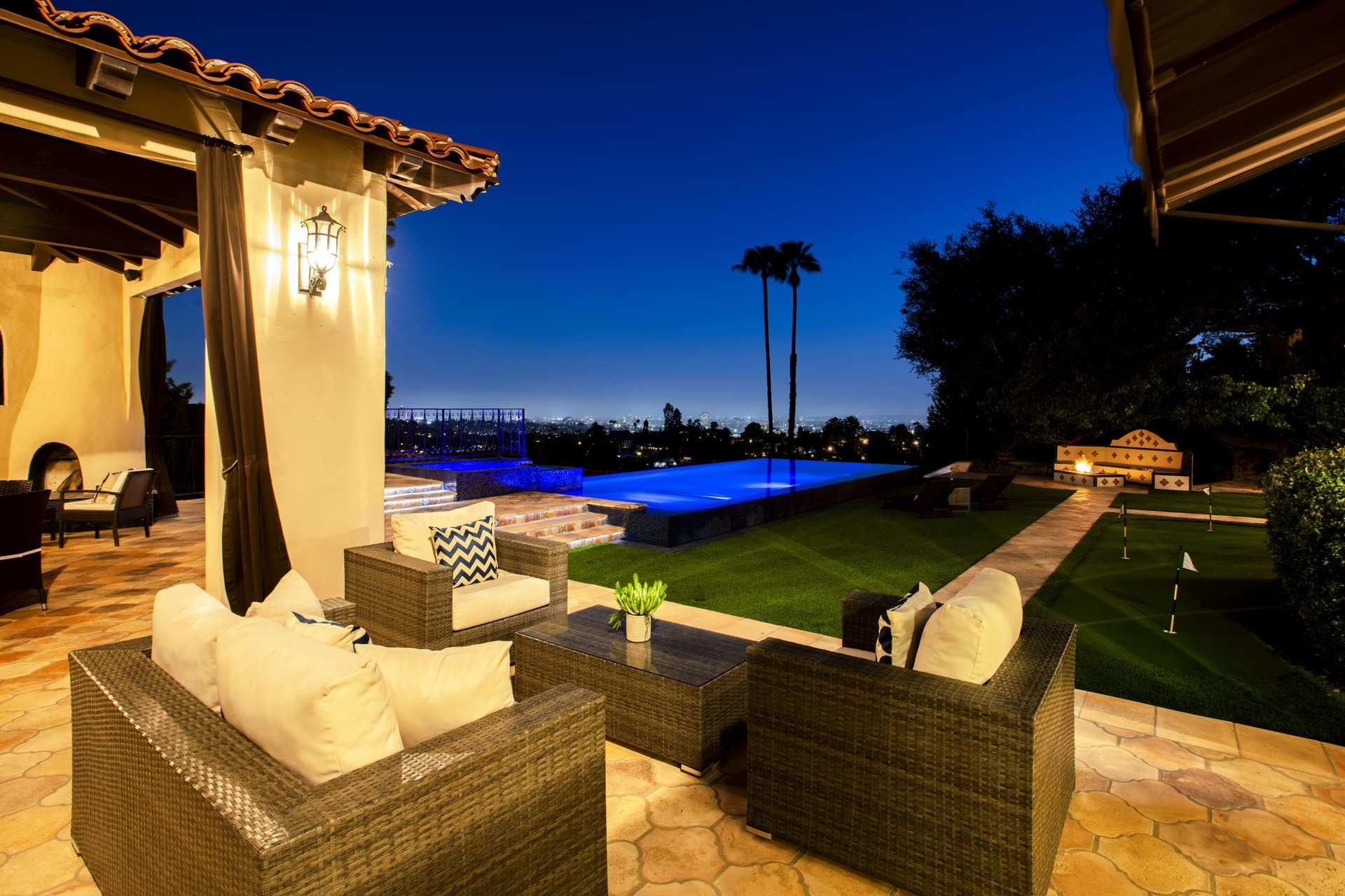 Outdoor patio with wicker seating beside a lit pool overlooking city lights at night.