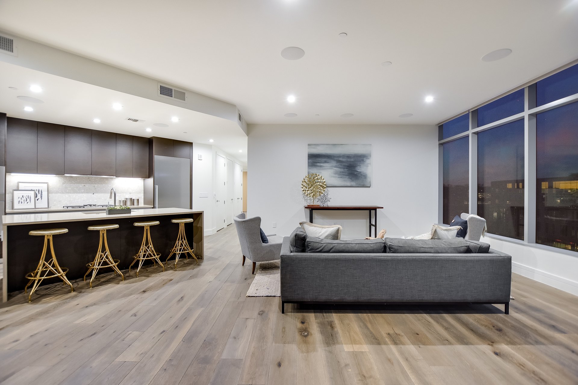 Open-plan condo living room with a breakfast bar, gray sofa, wood floors, and city views through tall windows.