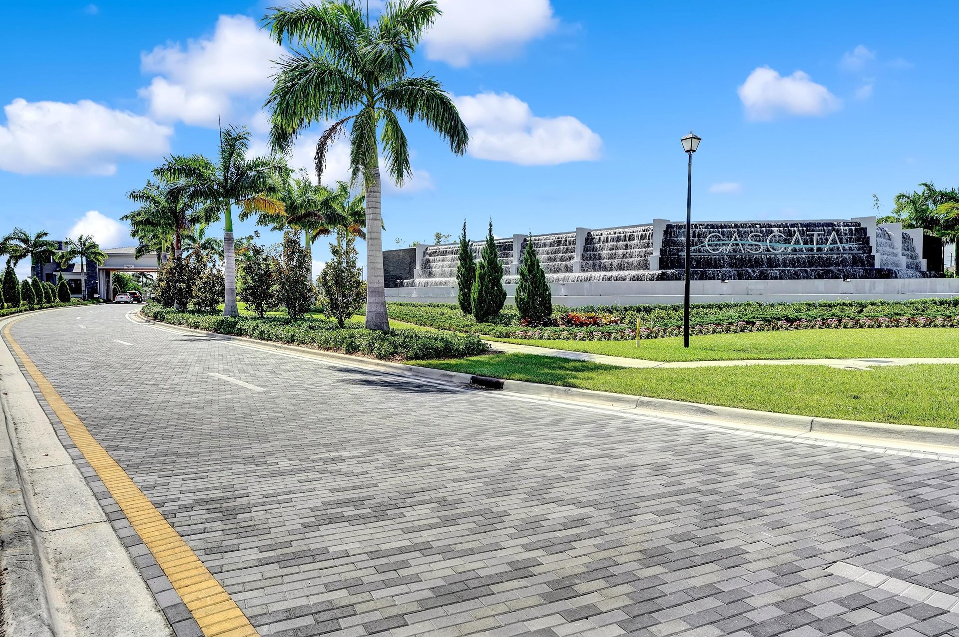 Cascata community entrance with a tiered waterfall feature, palm trees, and a curving paved road.