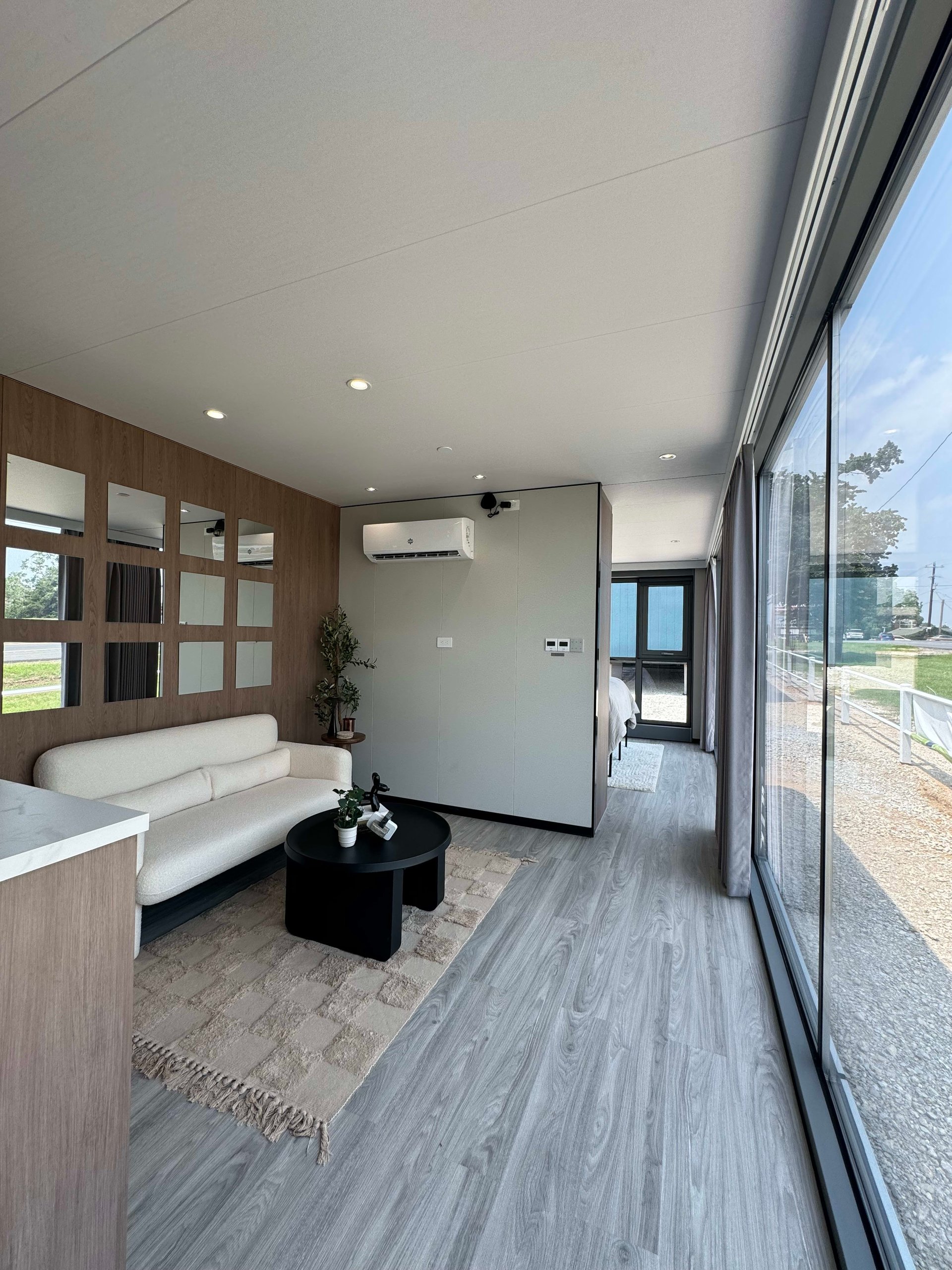 Tiny-home living room with a white sofa, mirrored wood wall, and floor-to-ceiling windows.