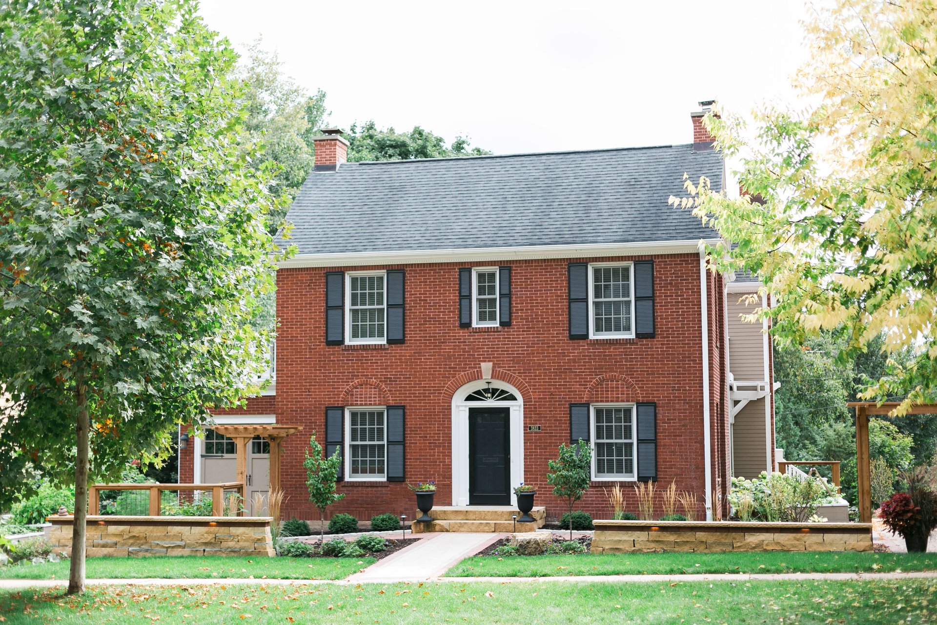 A red brick house with a black front door and black shutters, white trim, and a green front yard with flowers and shrubs.