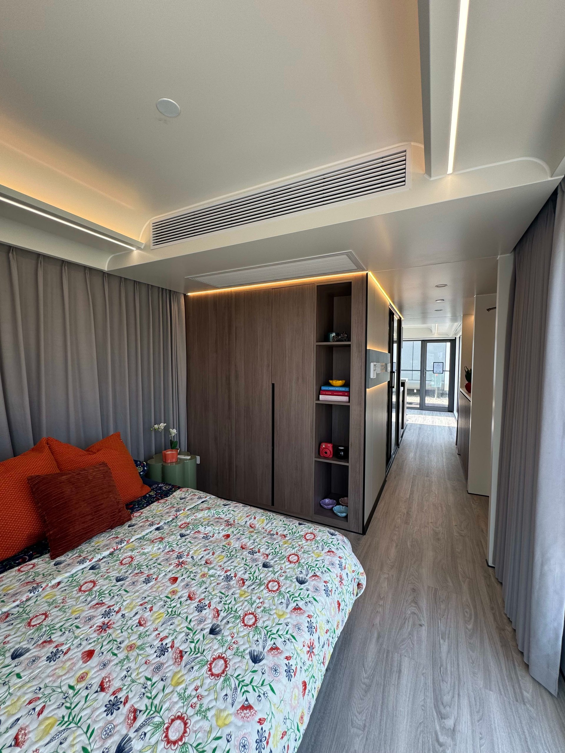 Tiny home bedroom with floral bedding, orange pillows, and built-in wood cabinetry beside a narrow hallway.