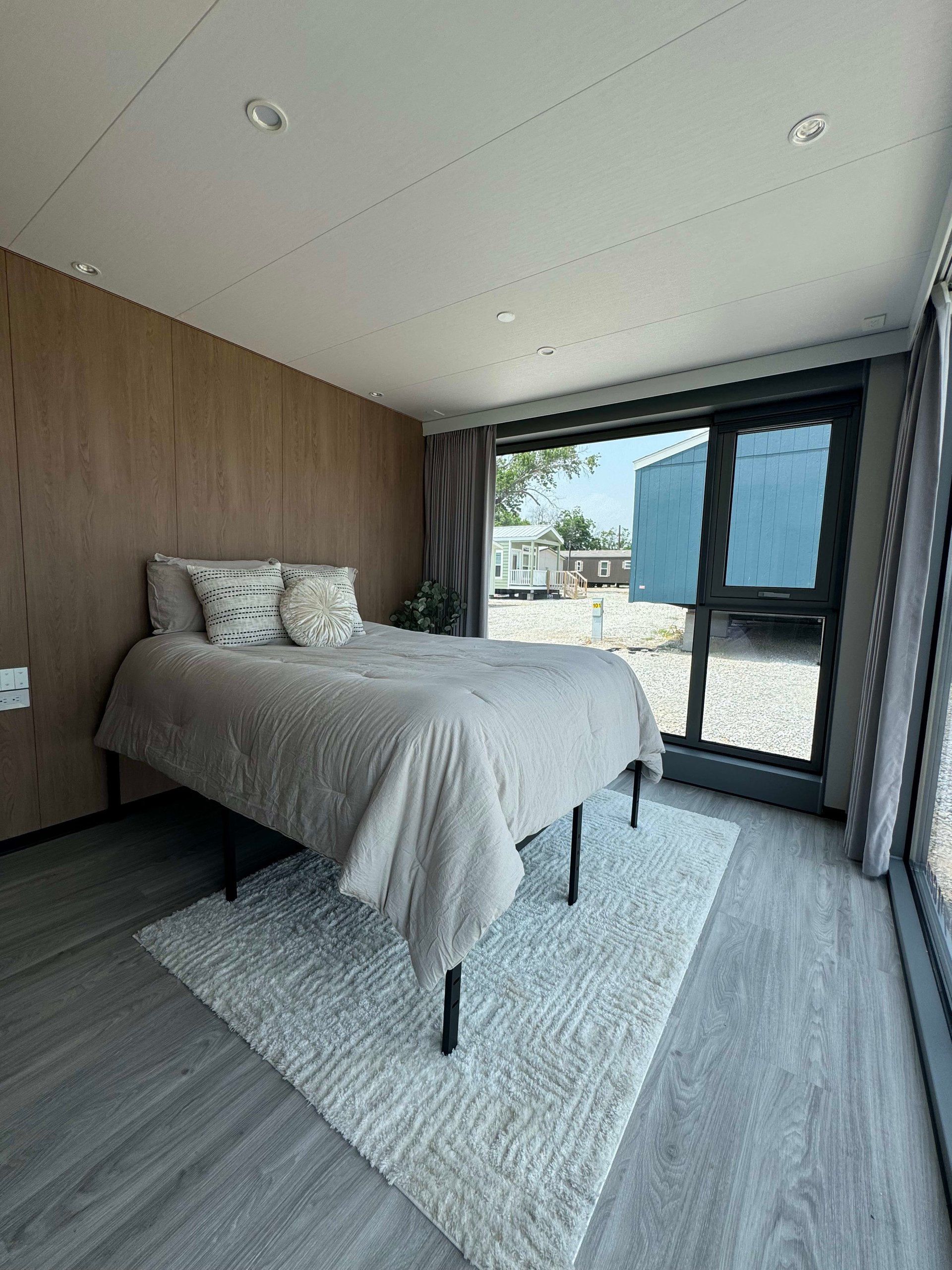 Tiny-home bedroom with a gray-covered bed, wood-paneled wall, and large windows.