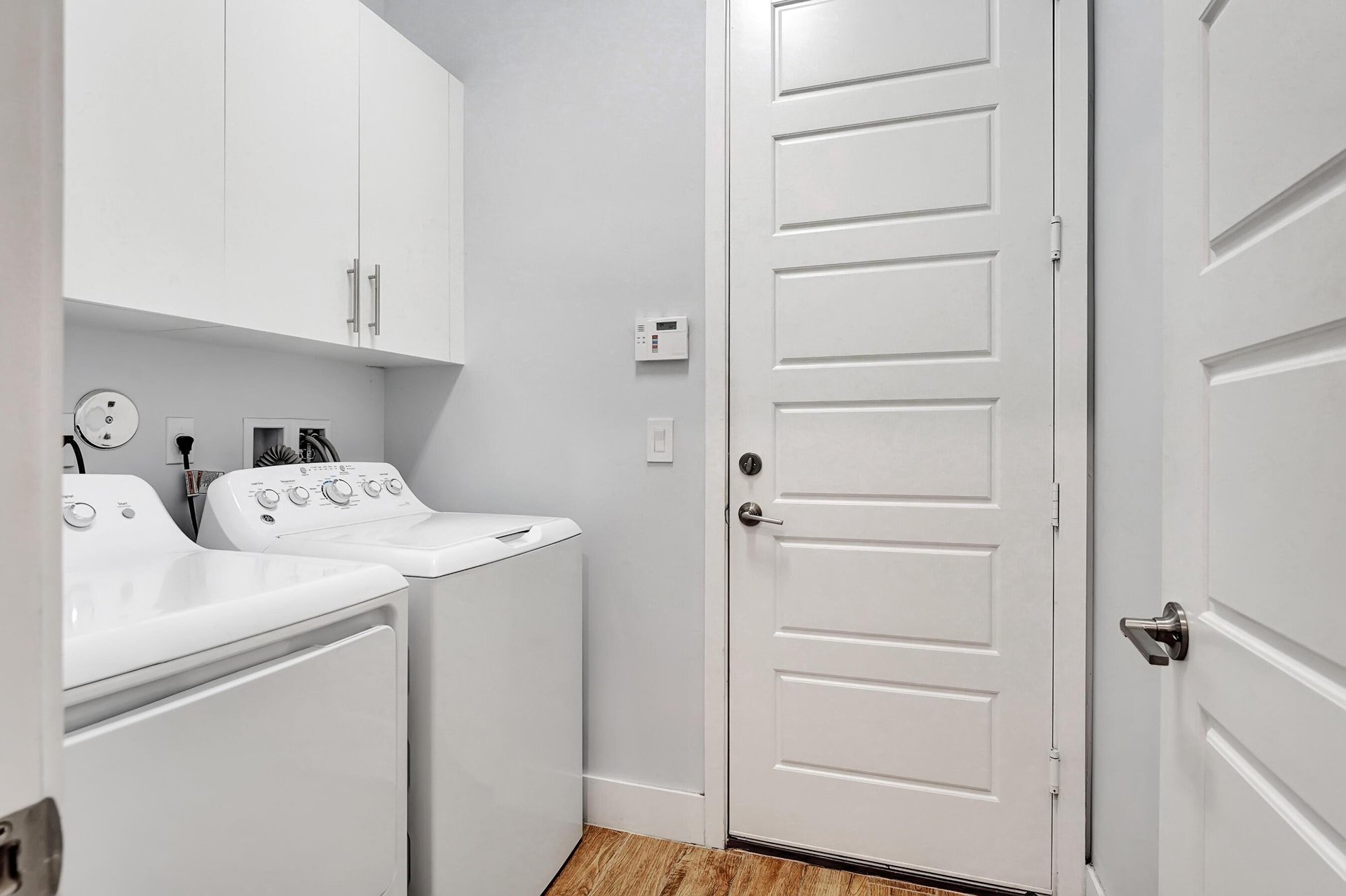 Laundry room with white washer and dryer beneath wall cabinets and wood-look flooring.