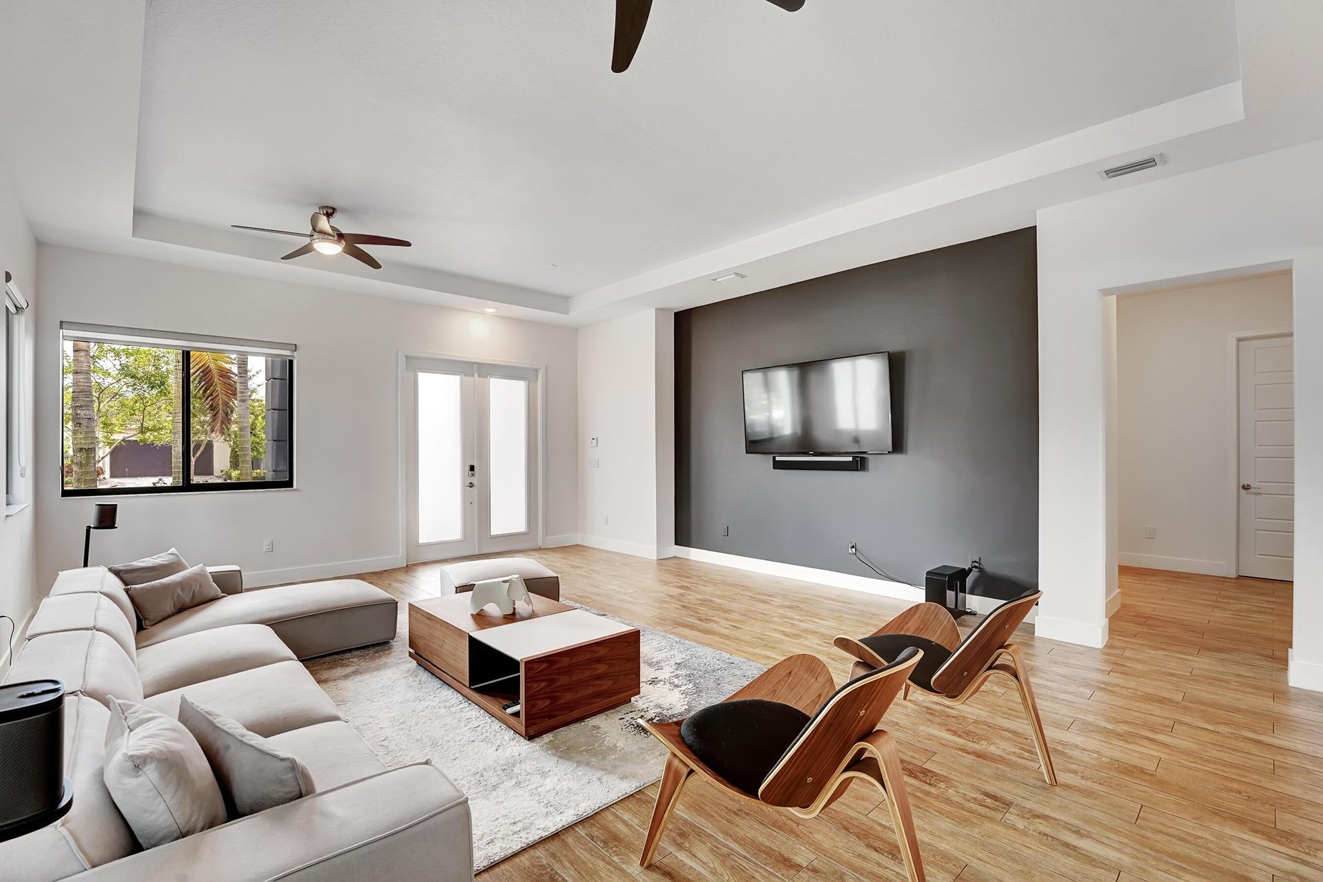 Furnished living room with a white sectional, wooden lounge chairs, and a television on a dark accent wall.