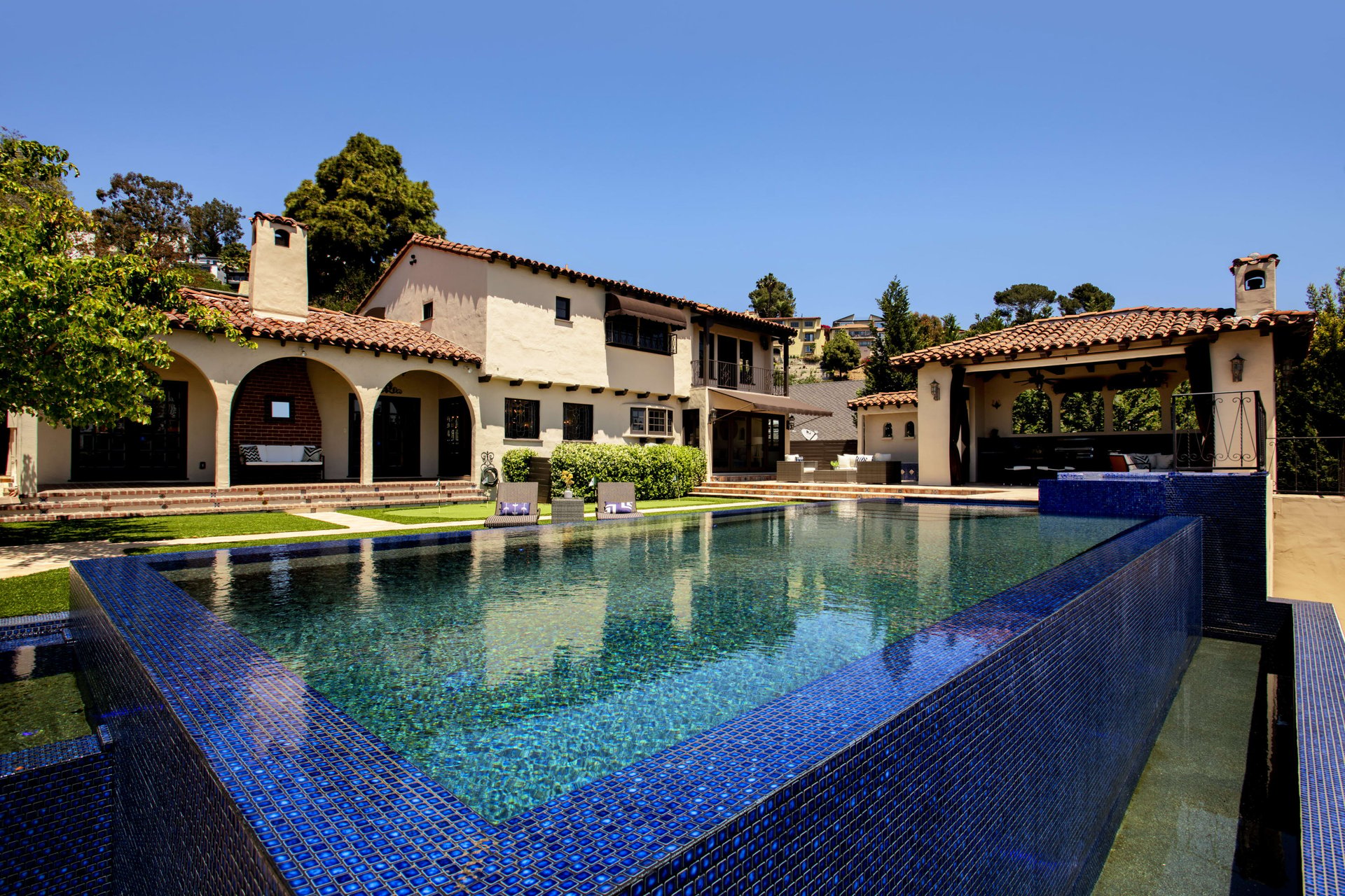 Spanish-style home behind a large blue-tiled pool and covered pavilion.
