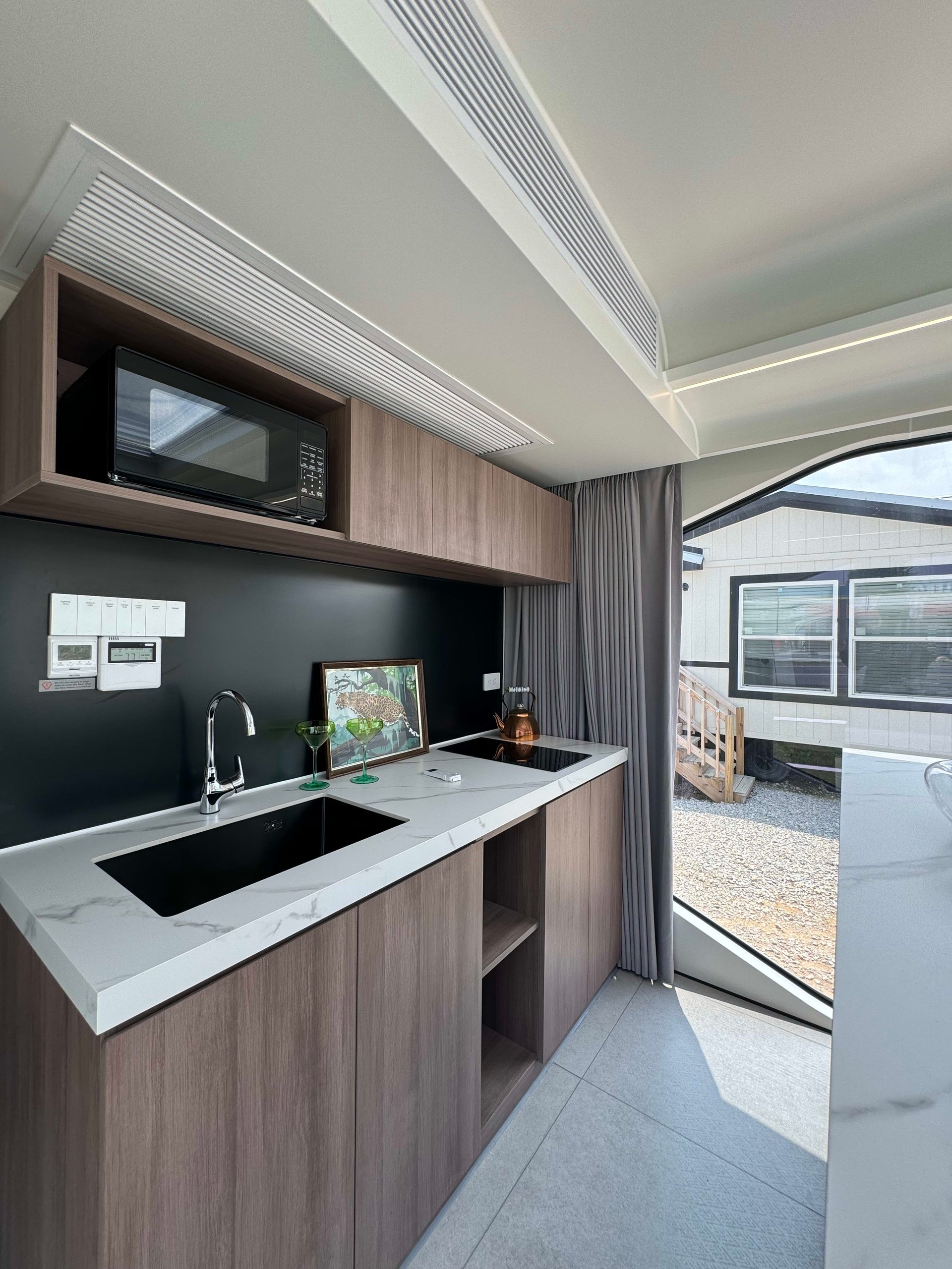 Tiny-home kitchen with wood cabinetry, a white counter, black sink and cooktop, and a large window.