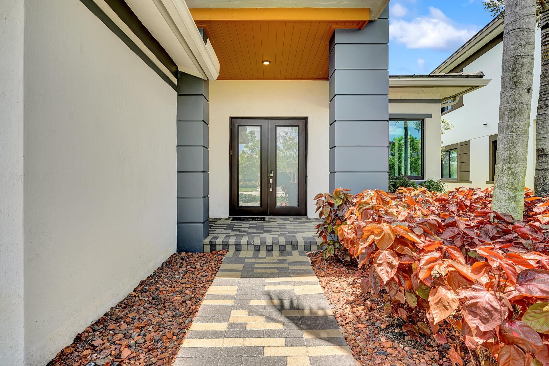 Modern home entrance with dark double doors, wood-paneled porch ceiling, and patterned paver walkway.