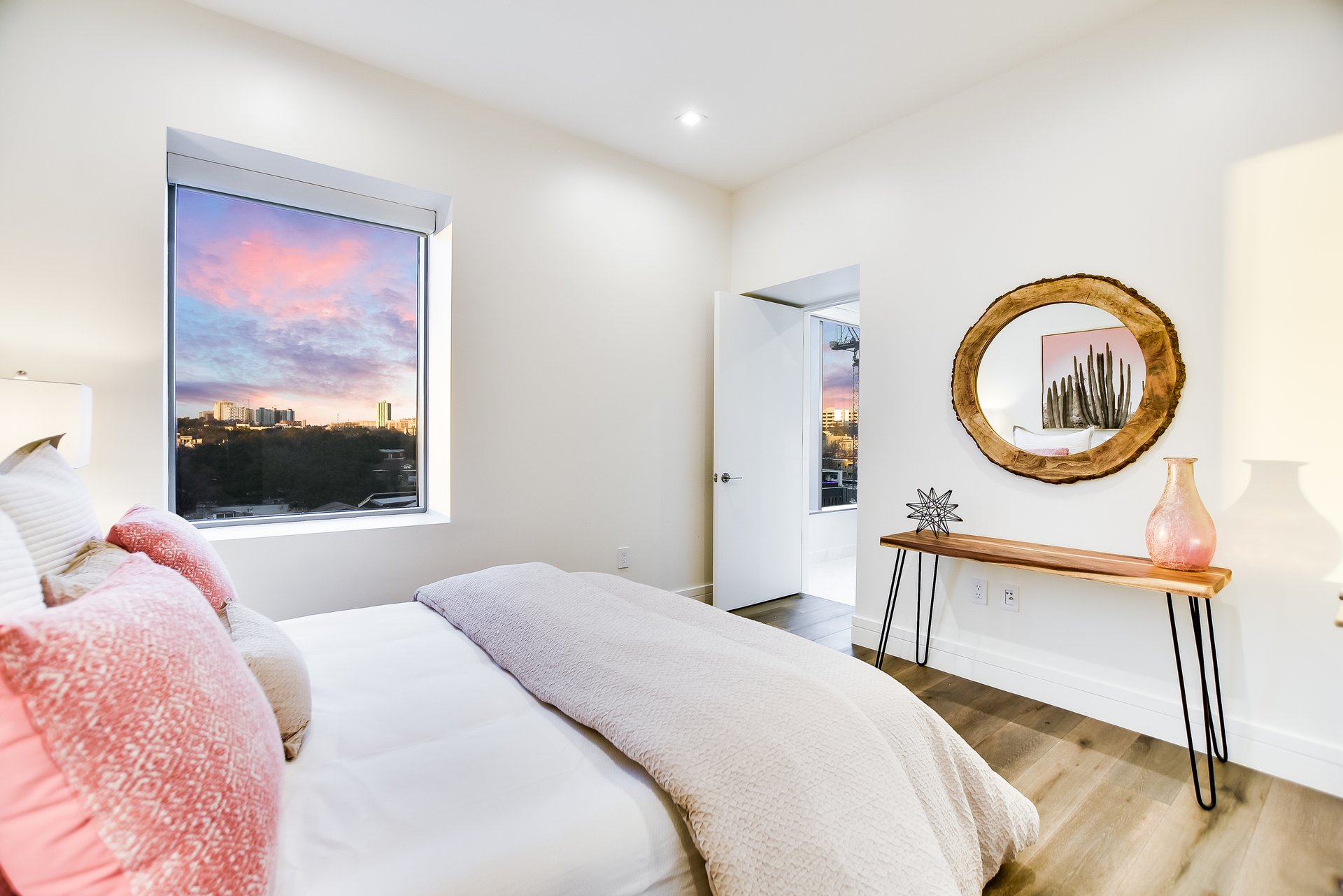 Bedroom with a large window overlooking trees and a city skyline at sunset.