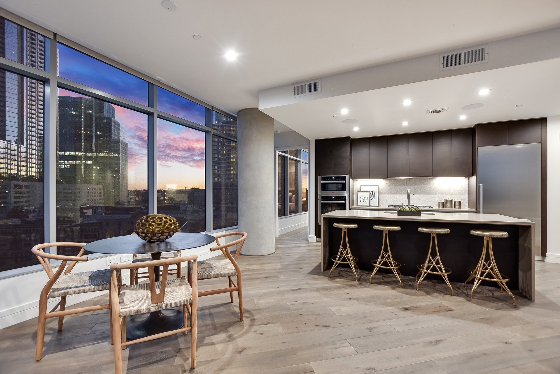 Furnished condo kitchen with a large island and floor-to-ceiling windows overlooking the skyline at sunset.