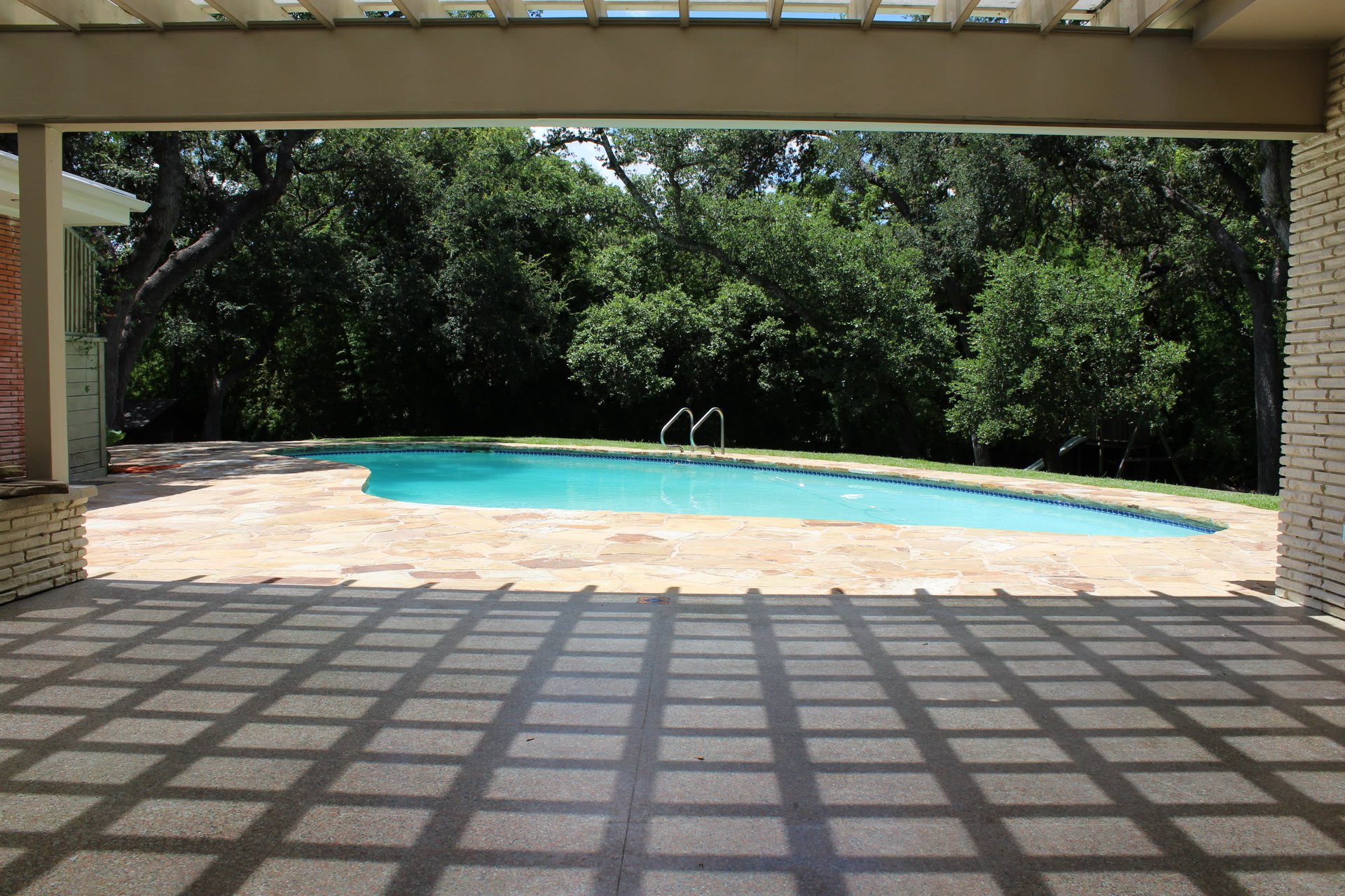 Swimming pool with a flagstone deck, viewed from a shaded covered patio against mature trees.