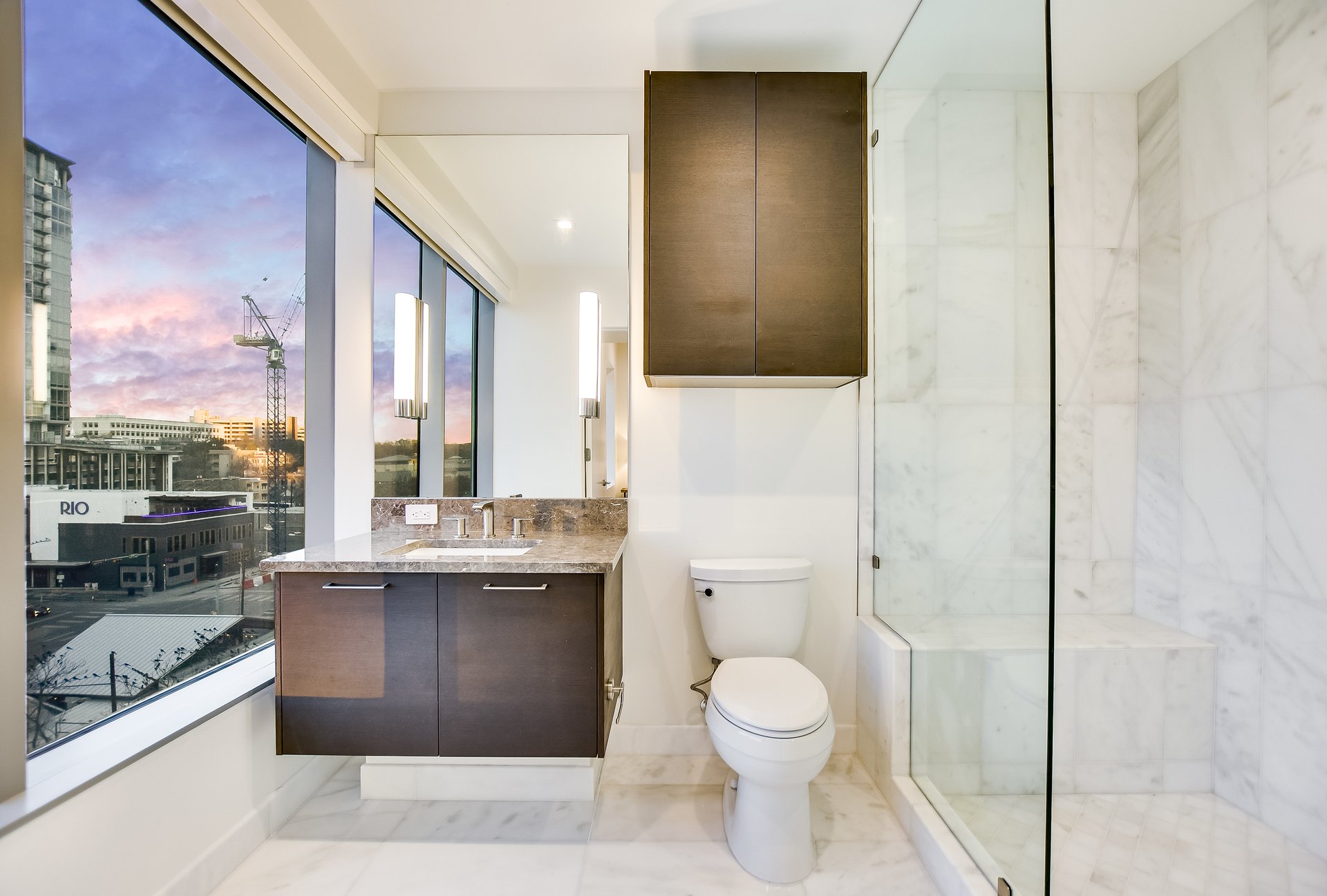 Modern bathroom with a marble shower and floor-to-ceiling window overlooking downtown at sunset.