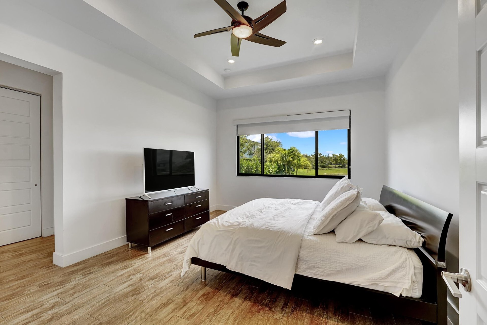 Bedroom with white bedding, a dark bed frame, ceiling fan, and window overlooking palm trees and lawn.