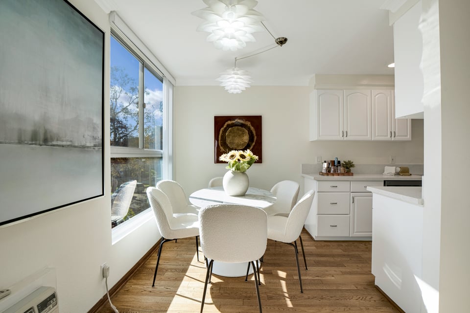Dining area with a round table beside large windows, adjoining a white kitchen.