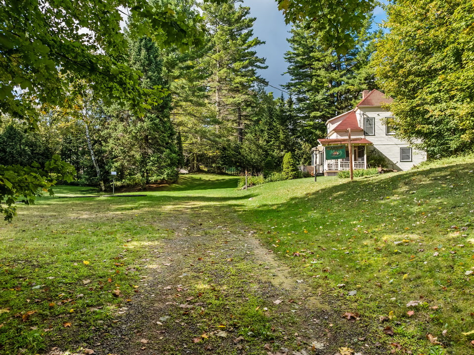 White two-story house with a red roof and covered porch amid mature trees and grassy grounds.