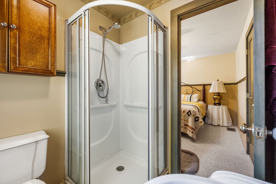 Bathroom with a glass-enclosed corner shower and wooden cabinet, opening onto a bedroom.