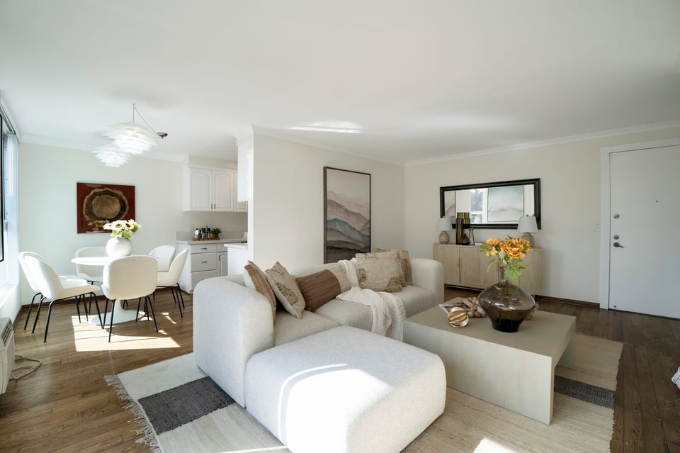 Staged living and dining area with a white sectional, neutral decor, and sunlight through large windows.