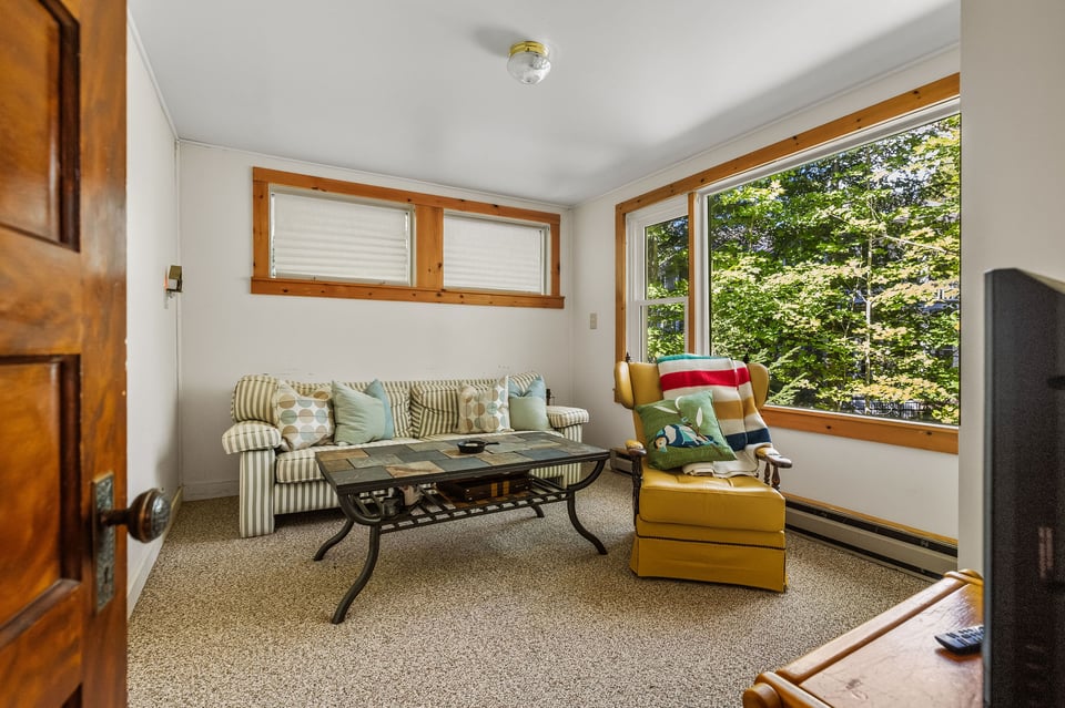 Living room with a striped sofa and yellow armchair beside large windows overlooking leafy trees.