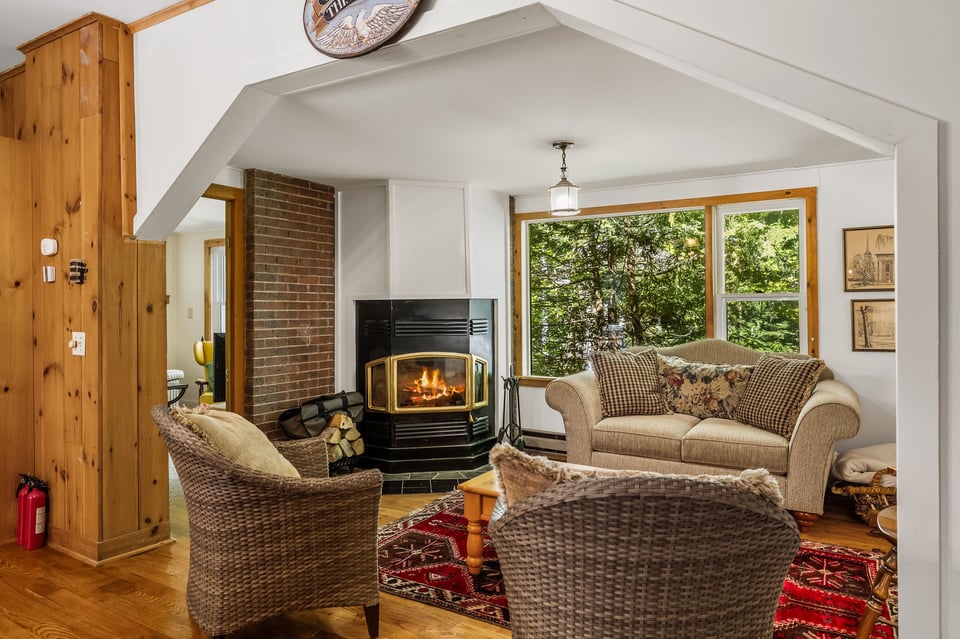 Living room with a lit fireplace, wicker seating, and large windows overlooking trees.