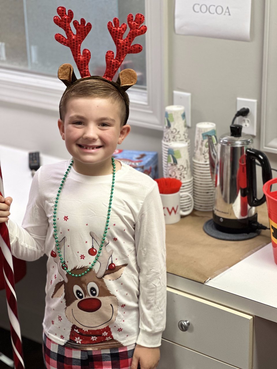 Child in reindeer antlers and a Christmas shirt stands beside a cocoa station.