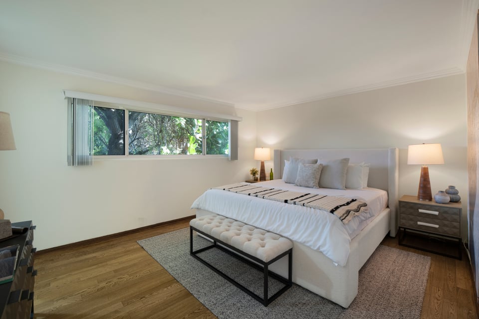 Bedroom with a large bed and upholstered bench beside a window overlooking leafy trees.