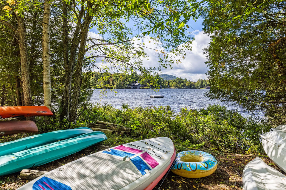 Kayaks and paddleboards rest beneath lakeside trees, facing a wooded shore across the lake.