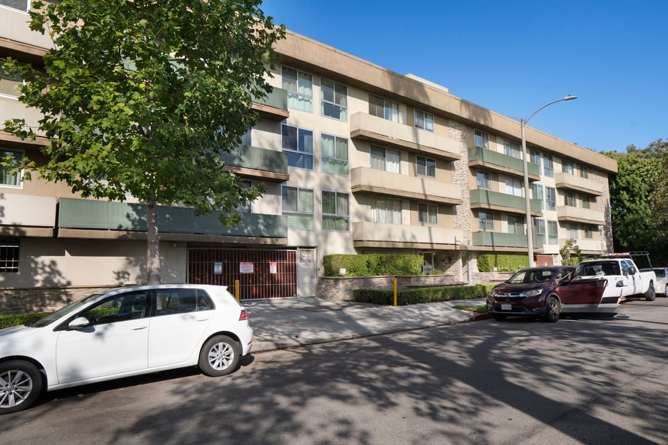 Multi-story apartment building with green balcony railings, partly screened by a leafy tree.