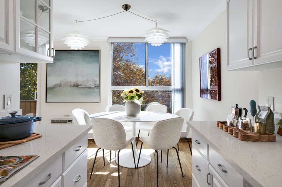 Bright kitchen with white cabinetry, a dining table, and large windows overlooking autumn trees.