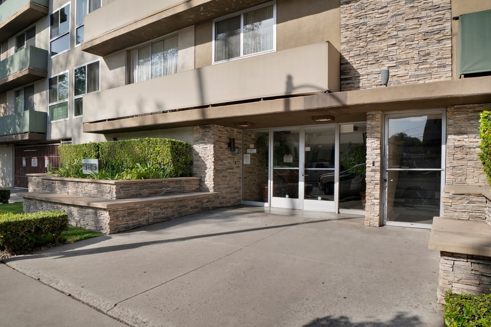 Apartment building entrance with glass doors, stone facade, and landscaped planter.
