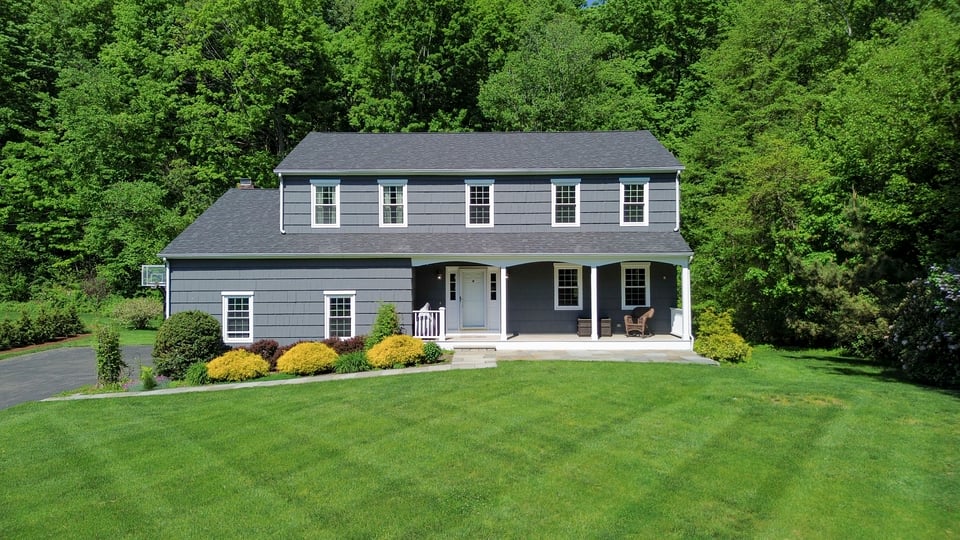 Gray two-story house with a covered porch and broad lawn, set against mature trees.