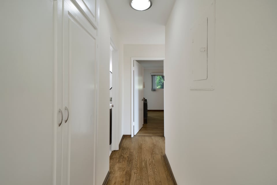 Apartment hallway with wood floors, white built-in cabinets, and an open doorway at the far end.