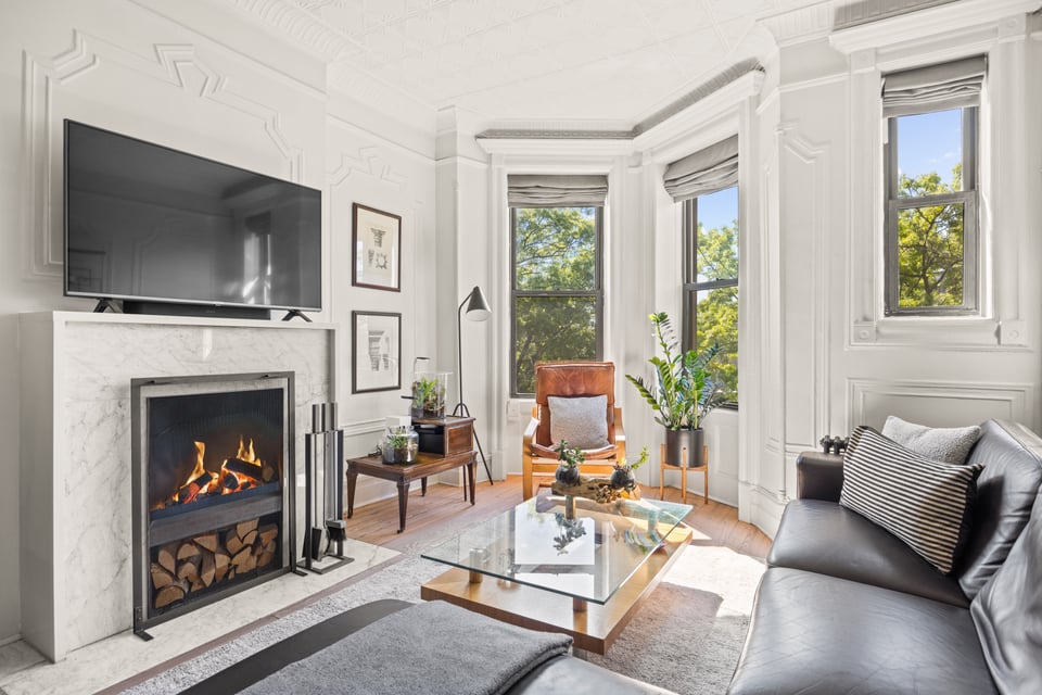 Staged living room with a marble fireplace, ornate moldings, and tall windows overlooking trees.