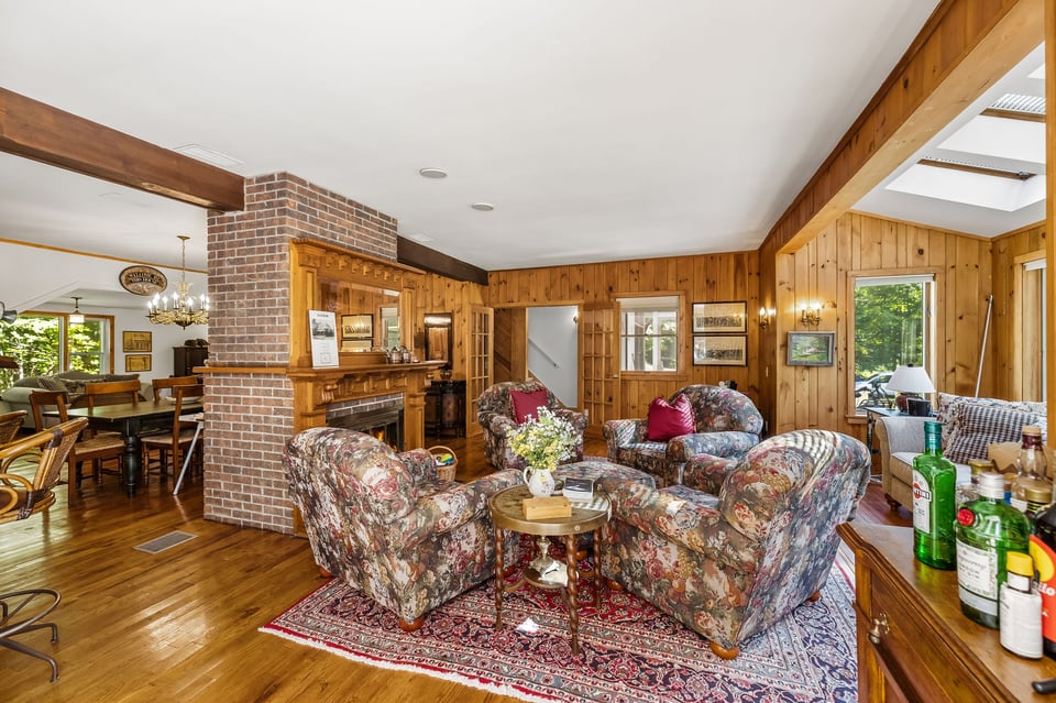 Wood-paneled living room with a brick fireplace, patterned seating, and skylights.