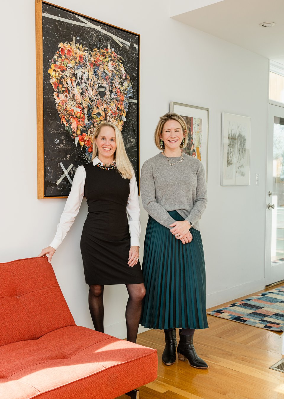 Two women stand together in an office beside a red-orange sofa and colorful framed artwork.