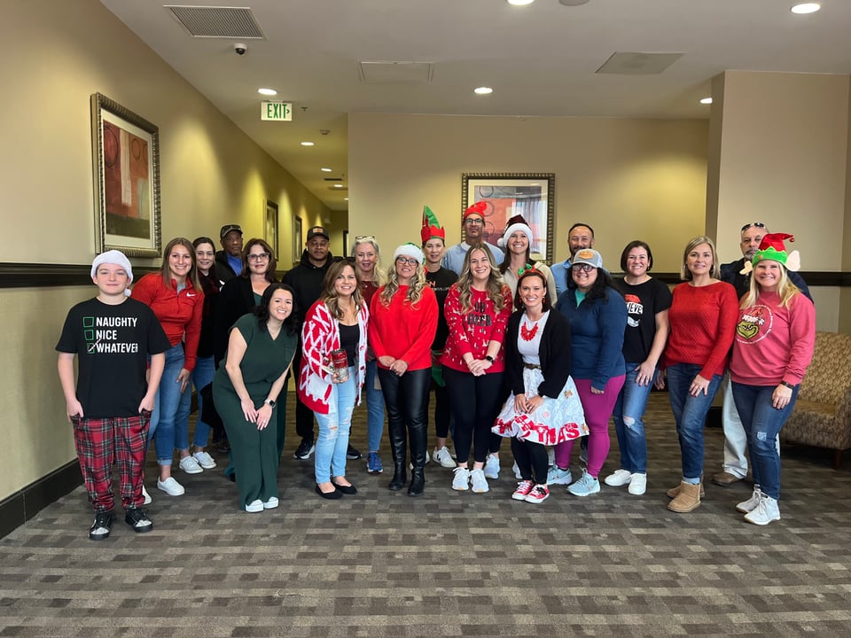 A group poses in Christmas sweaters and Santa and elf hats in a hallway.
