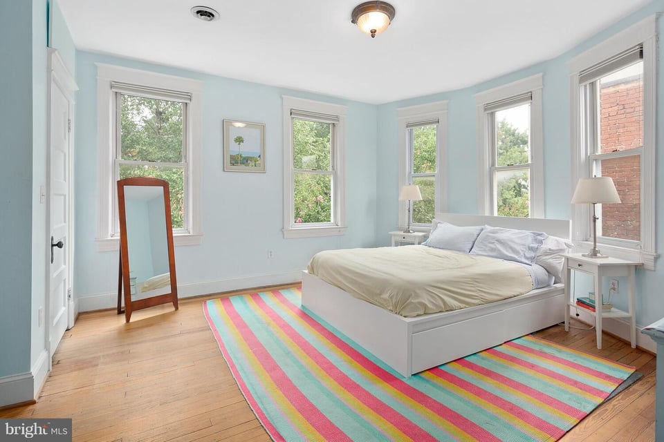 Bedroom with a white bed, pale blue walls, hardwood floors, and a colorful striped rug.