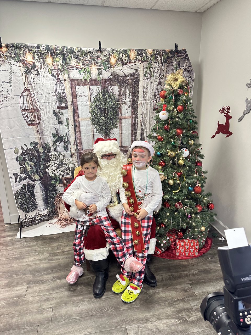 Two children in festive pajamas sit with Santa beside a decorated Christmas tree.