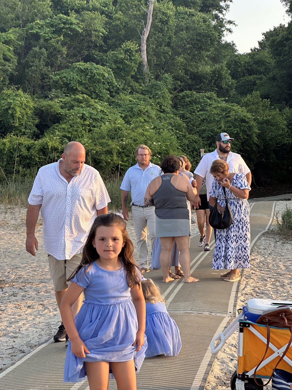 A girl in a blue dress stands on a sandy beach boardwalk as a group walks behind her.
