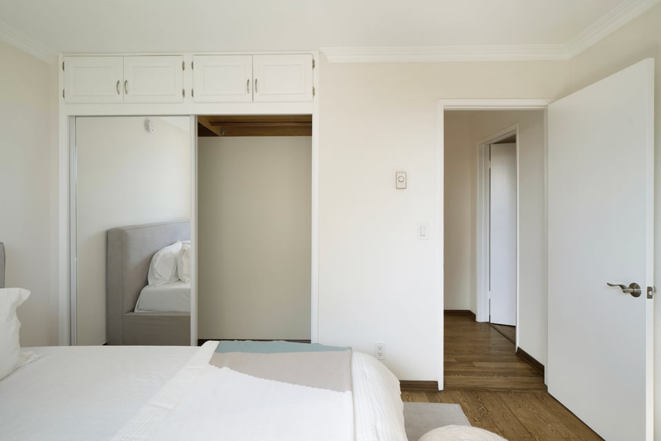 Bedroom with mirrored closet doors and overhead cabinets, looking toward an open doorway and wood-floored hall.