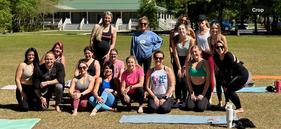 A group of adults pose on a lawn beside yoga mats, with a white building behind them.