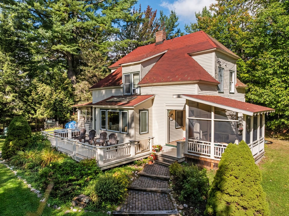 White house with a red roof, screened porch, and deck surrounded by trees and gardens.