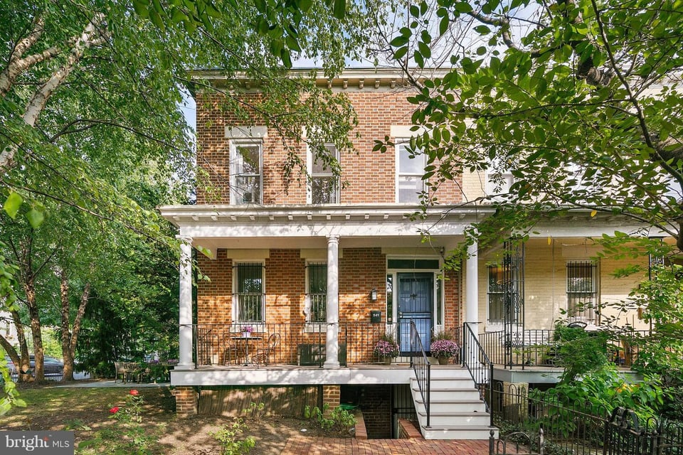 Red-brick rowhouse with a white-columned porch, blue door, and leafy trees framing the front yard.