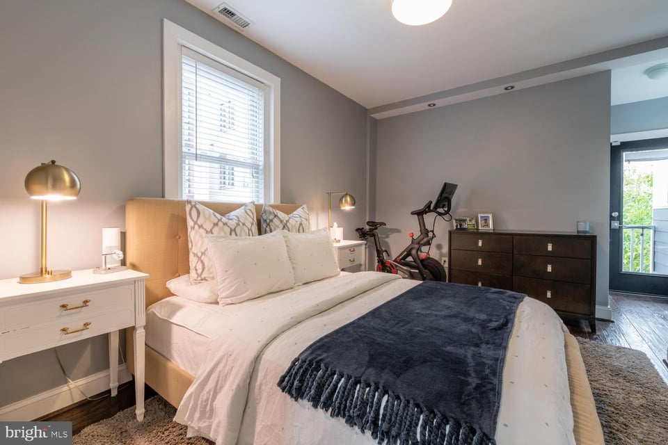 Furnished bedroom with a white bed, navy throw, exercise bike, and balcony door.