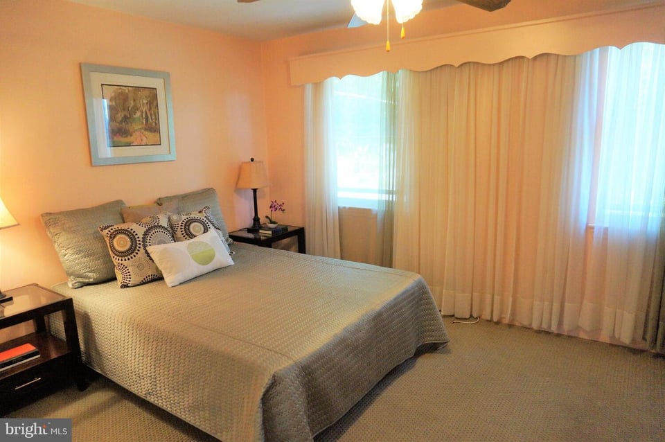 Furnished bedroom with a large beige-quilted bed and broad curtained windows.