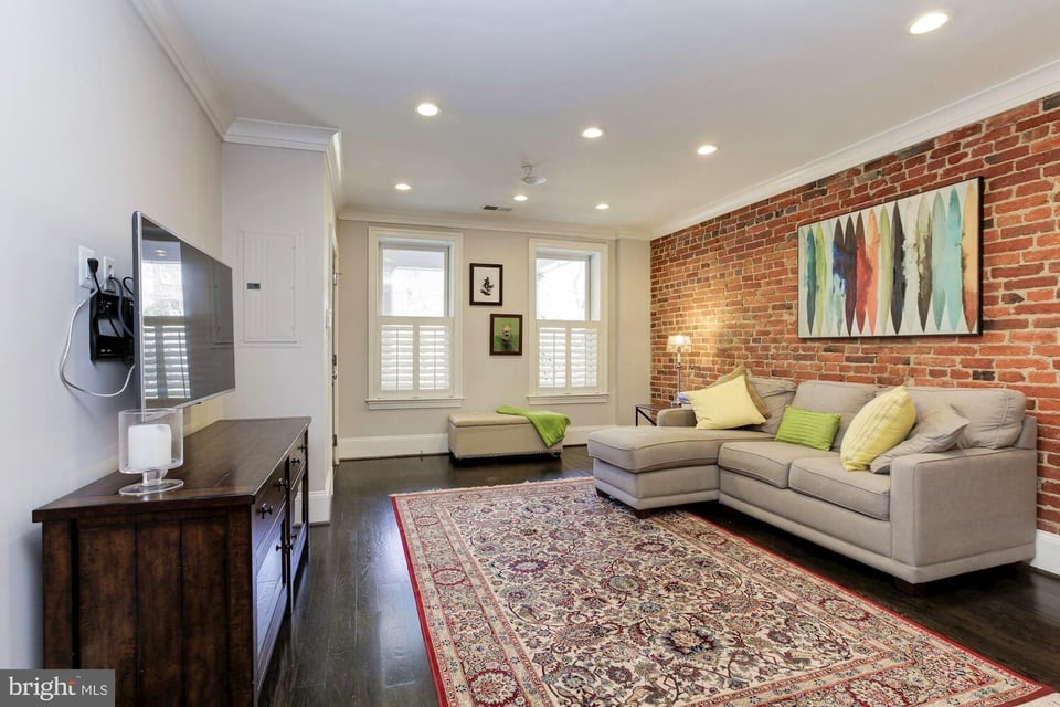 Living room with a gray sectional, exposed brick wall, patterned rug, and shuttered windows.