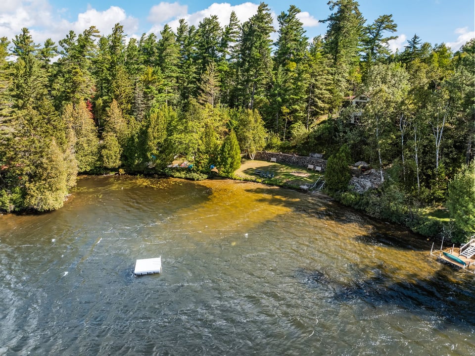 Aerial view of a wooded waterfront property with a floating dock and landscaped shoreline lawn.