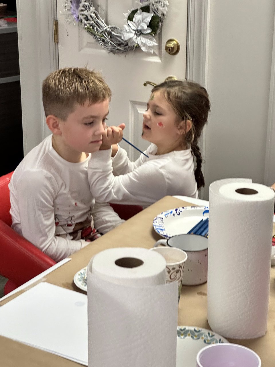 Two children share an art activity at a table, with paper towels and plates nearby.