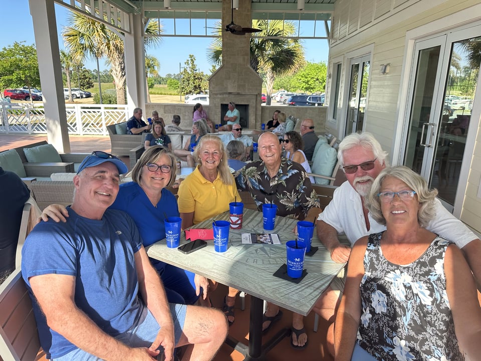 Adults gather around a table with blue cups at a sunny covered patio event.