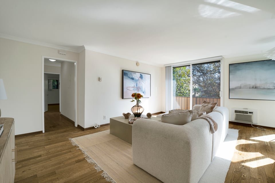 Staged living room with a cream sofa, wood floors, and sliding glass doors to a tree-lined balcony.